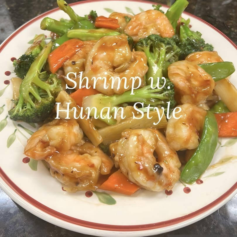 Hunan-style shrimp with broccoli, carrots, and snow peas in a savory sauce on a white plate.